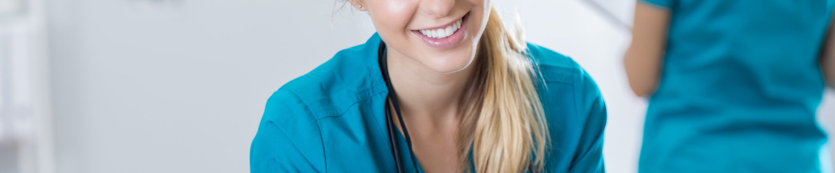 Smiling Caucasian female nurse uses a smart phone while working in a doctor's office or hospital. She is also using a laptop. A nurse is working in the background.