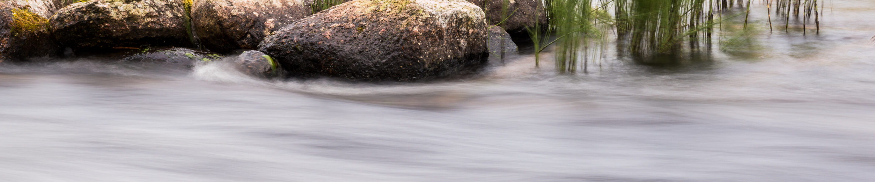 Long exposure shot of streaming river in Finland with stones and sedge vegetation in the background.